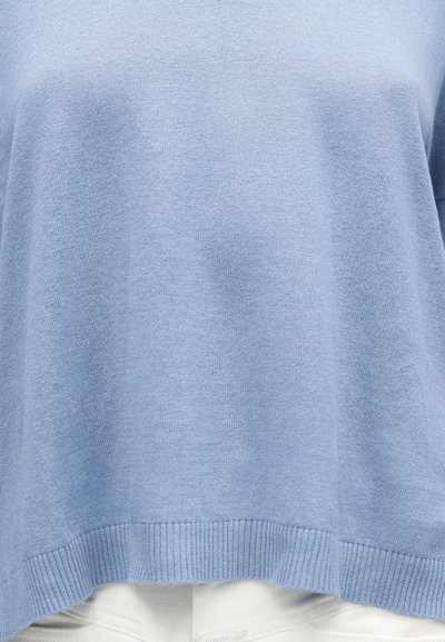 Ανοιχτό μπλε πλεκτό πουλόβερ με ριμπ τελείωμα, φορεμένο πάνω από λευκό παντελόνι, κομμένο στη μέση, που δείχνει την υφή και τις λεπτομέρειες του υφάσματος.