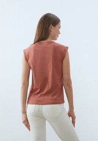 Top sin mangas de color óxido con ribete de encaje en los hombros, hecho de una tela suave, combinado con jeans blancos, visto desde la parte trasera.