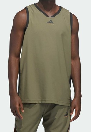 Homme portant un maillot de basket sans manches vert olive et un short assorti avec des logos Adidas noirs et orange sur fond gris clair.