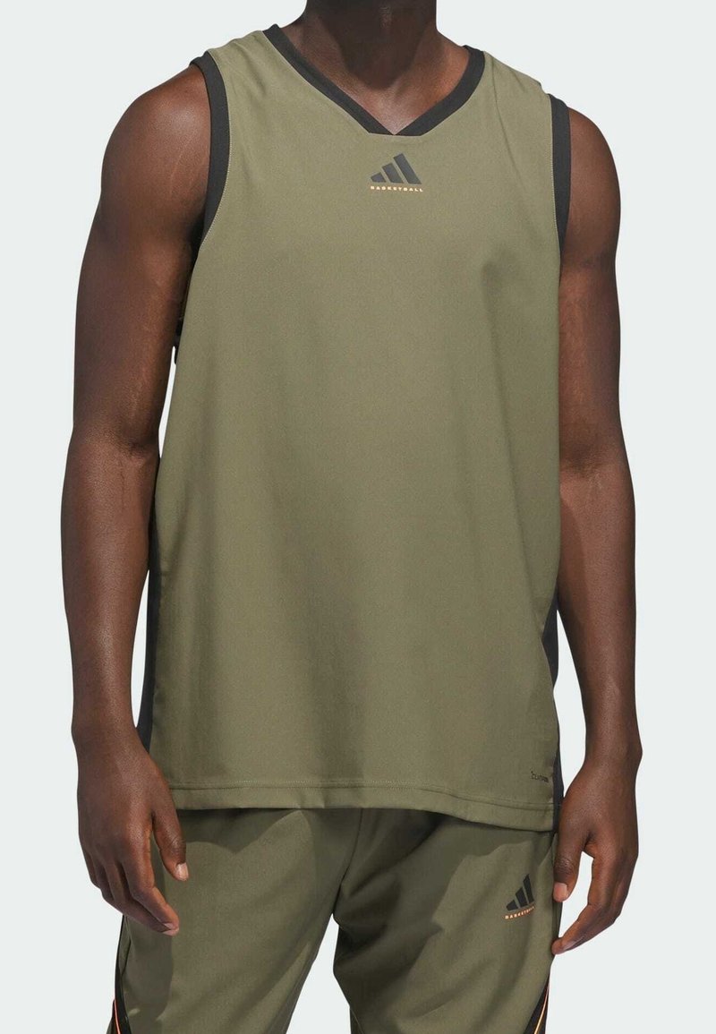 Homme portant un maillot de basket sans manches vert olive et un short assorti avec des logos Adidas noirs et orange sur fond gris clair.