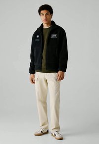 Juoda fliso striukė su užtrauktuku priekyje, pleistrų detalėmis ir laisvu kirpimu, derinama su smėlio spalvos cargo kelnėmis ir baltais sportiniais bateliais.