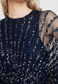 Gros plan sur le haut du torse d'une femme portant une robe noire avec des sequins noirs en diagonale sur les manches en tulle transparent et le corsage.