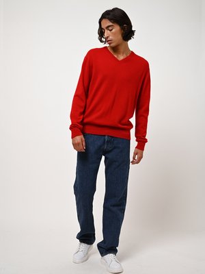 Rode gebreide v-hals trui, losse pasvorm, gedragen met donkerblauwe rechte jeans en witte sneakers. Glad textuur, casual ontwerp.