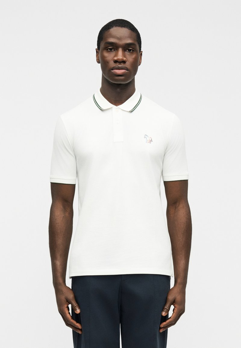 Νεαρός άνδρας που στέκεται με το πρόσωπο προς τα εμπρός, φοράει λευκό πόλο μπλουζάκι με ριγωτό γιακά και σκούρο παντελόνι, σε απλό λευκό φόντο.