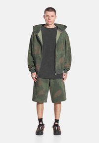 Zielona bluza z kapturem na zamek w kamuflażu, szara koszulka, zielone krótkie spodenki w kamuflażu, czarne skarpetki za kostkę i czarne trampki, prezentujące swobodny krój i casualowy styl.