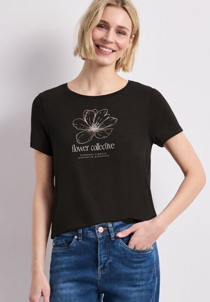 Uśmiechnięta kobieta z krótkimi blond włosami, ubrana w czarną koszulkę z szkicem kwiatu i napisem „flower collective”, w połączeniu z niebieskimi jeansami.