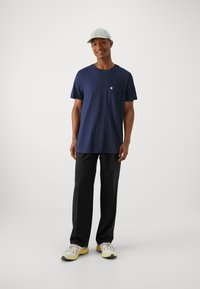 T-shirt en coton bleu marine avec une poche au niveau de la poitrine, associé à un pantalon noir et des baskets claires avec un design texturé et des accents jaunes.