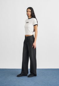 Νεαρή γυναίκα με μακριά σκούρα μαλλιά, ντυμένη με άσπρο κοντό τοπ με μαύρη λεπτομέρεια, φαρδιά μαύρα τζιν και μαύρα αθλητικά παπούτσια, στέκεται σε μπλε χαλί.