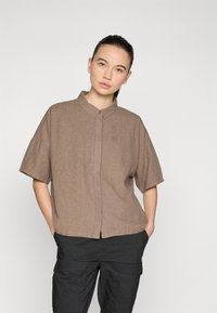 Bruine, korte mouw knoopshirt van een lichte stof met een subtiele textuur. Heeft een kraag en een klein geborduurd detail.