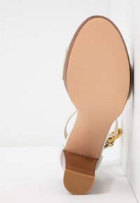 Sandale à talon avec une tige en cuir beige, une semelle en caoutchouc texturé et un accent en bois clair sur le talon. Détail de boucle dorée inclus.