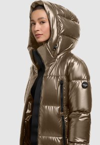 Woman wearing metallic bronze puffer jacket with large hood, black zipper, and brand patch on left sleeve, shown from the chest up.