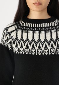 Svart stickad tröja med en vit mönstrad okdel som har geometriska mönster. Mjuk textur, rund hals och medelviktsmaterial.