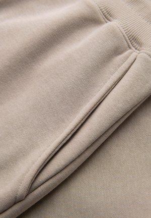 Primo piano del tessuto beige dei pantaloni della tuta che mostra una tasca laterale e le cuciture della vita elastica.