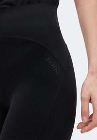 Κοντινό πλάνο ενός ατόμου που φοράει μαύρο κολάν με ψηλή μέση, ορατές ραφές και το λογότυπο «BORG» στην περιοχή των γοφών.