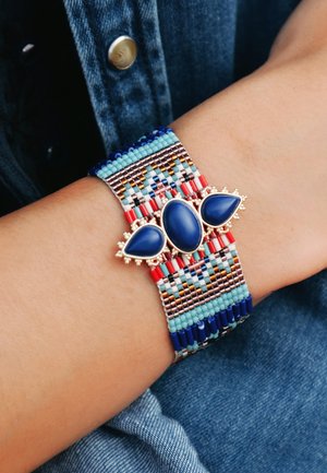 Perlenarmband mit blauen Steinen, goldenen Akzenten und komplizierten Mustern aus roten, blauen und türkisfarbenen Perlen, ergänzt durch Fransen-details.
