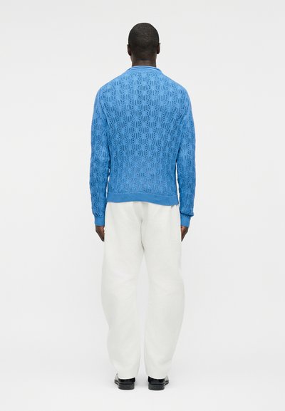 Άνδρας που στέκεται, γυρισμένος από την πλάτη, φοράει μπλε πουλόβερ με μακριά μανίκια και σχέδια, άσπρο φαρδύ παντελόνι και μαύρα παπούτσια μπροστά σε απλό φόντο.