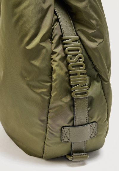 Γωνία μαξιλαρωμένης τσάντας σε ελιά πράσινο με κάθετο δερμάτινο λουράκι που φέρει μεγάλα γράμματα της μάρκας MOSCHINO στο ίδιο χρώμα.