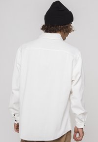 Uomo che indossa una camicia a maniche lunghe bianca e un berretto nero, di spalle, su uno sfondo grigio chiaro uniforme.