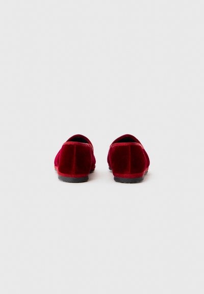 Červené zamatové slip-on topánky s okrúhlym tvarom, čiernymi gumovými podrážkami a hladkou textúrou. Minimalistický dizajn bez viditeľných kovania alebo akcentov.
