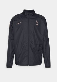 Svart Nike-jacka med dragkedja, hög krage och Spurs-logotyp. Har elastiska ärmslut och sidofickor, tillverkad i lätt material.
