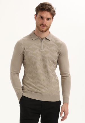Uomo che indossa un maglione polo beige a maniche lunghe con un motivo geometrico testurizzato e pantaloni neri, in piedi contro uno sfondo bianco uniforme.