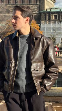 Rjava usnjena jakna z zadrgo spredaj in dvema žepoma na prsih, nošena preko sivega puloverja. Model stoji na prostem pred zgodovinsko kuliso.