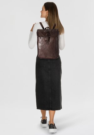 Zaino in pelle marrone con spalline regolabili, chiusura a pattina e design minimalista. Il modello indossa una gonna nera e sneakers.