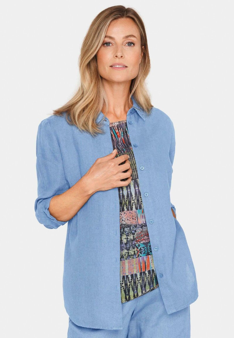 Helles blaues Button-Up-Hemd mit lockerer Passform, das hochgerollte Ärmel und eine gemusterte Innenlage mit verschiedenen Farben und Texturen aufweist.