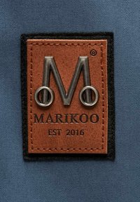 Brūna ādas plāksne ar izspiestu sudraba "M" logo, tekstu "MARIKOO" un "EST 2016", apkārt melna šuvuma apmale, uz zila auduma fona.
