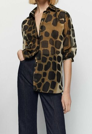 Woman wearing a brown and black spotted open-collar blouse with rolled-up sleeves and dark high-waisted pants, hand in pocket.