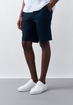 Uomo con pantaloncini blu navy, scarpe da ginnastica bianche e camicia azzurra, con una mano in tasca, in piedi su pavimento grigio contro sfondo bianco.