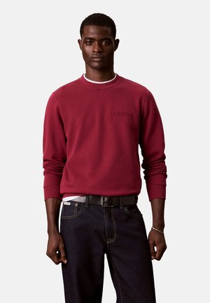 Hombre con sudadera Calvin Klein de color rojo oscuro, cinturón negro y vaqueros oscuros, de pie frente a un fondo claro y simple.