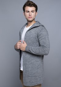 Grå strikket cardigan med hætte, der har lange ærmer, en struktureret overflade og åben front design. Sammen med en hvid skjorte inderst.