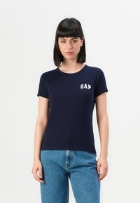 Giovane donna con capelli lisci scuri, che indossa una maglietta navy GAP e jeans blu, in piedi contro uno sfondo bianco semplice.