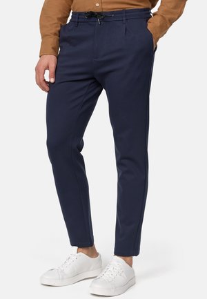 Homme portant un pantalon bleu marine fuselé avec taille élastique noire à cordon, chemise marron et baskets blanches, les mains dans les poches, debout devant un fond blanc.