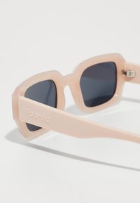 Roze prozirne sunčane naočale sa četvrtastim staklima i tamno obojenim staklima. "Polaroid" logo s strane, sa glatkom plastičnom teksturom.