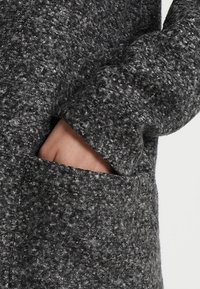 Grijs getextureerde jas met een zachte, wolachtige stof. Voorzien van een zijzak en losvallende mouwen. Het oppervlak heeft een gespikkeld patroon.