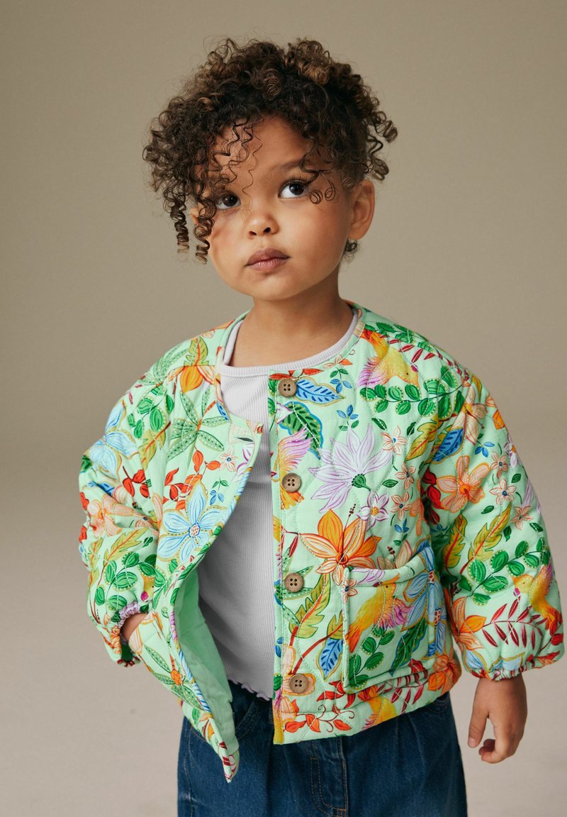 Chaqueta bomber acolchada en verde pastel con patrones florales multicolores, con cierres de botones y bolsillos laterales. Llevada sobre una camiseta gris.