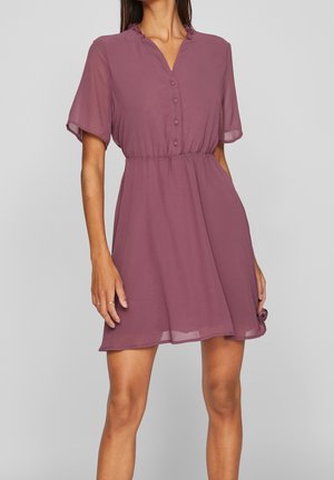 Kvinde iført en kortærmet lavendelfarvet kjole med indsnævret talje, knaplukning foran og flæsekant ved halsudskæringen, stående mod en ensfarvet baggrund.