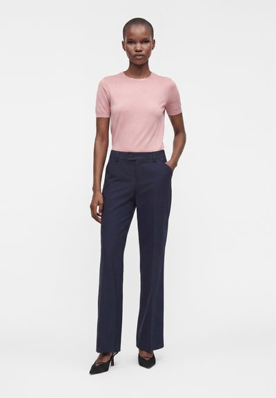 Femme debout portant un haut rose ajusté à manches courtes, un pantalon droit bleu marine et des chaussures à talons noirs, une main dans la poche.