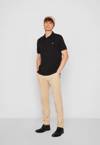 Juoda polo marškinėliai trumpomis rankovėmis, smėlio spalvos chino kelnės ir juodi ankletiniai batai. Komplektą papildo šviesiai smėlio spalvos kepuraitė, o marškinėliuose matomas mažas logotipas.