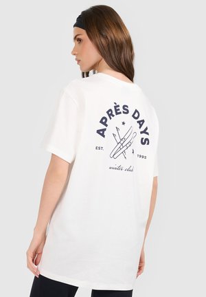 Kobieta w białej koszulce z granatowymi nartami i napisem "APRÈS DAYS winter club est. 1995" na plecach.