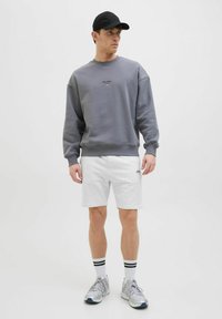 Sudadera gris con puños acanalados, que presenta un pequeño logo. Combinada con pantalones cortos blancos y zapatillas grises. Una gorra negra y calcetas a rayas completan el look.