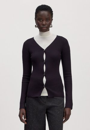Femme portant un cardigan noir côtelé avec des ouvertures pour boutons sur un col roulé blanc, associé à un pantalon texturé foncé et des boucles d’oreilles anneaux.