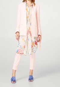 Kobieta ubrana w jasnoróżową marynarkę i spodnie z długą kwiecistą koszulą oraz niebieskimi, otwartymi klapkami na obcasie, stojąca na jednolitym tle.