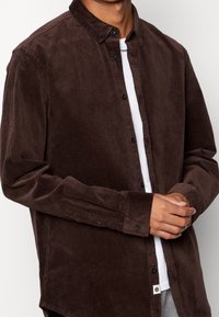 Hnědá manšestrová košile na knoflíky s rozloženým límcem, černými knoflíky a dlouhými rukávy. Pod ní je vidět bílá spodní košile. Texturovaná látka.