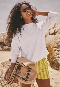 Weißes Rundhals-Sweatshirt, gelbe gestreifte Shorts und eine Strohtasche mit einem dekorativen Muster, vor einem Hintergrund aus feinem Sandstrand.