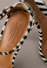 Tige en tissu rayé noir et blanc avec une semelle intérieure en cuir marron, présentant un logo "Signature" embossé et des détails de taille.