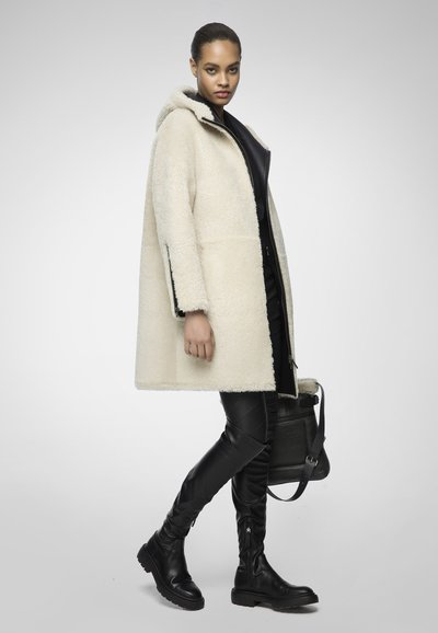 Donna che indossa un cappotto con cappuccio in shearling crema, pantaloni di pelle nera, stivali neri al ginocchio e tiene in mano una borsa in shearling scuro.