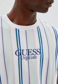 Camiseta de manga corta a rayas en blanco, con tonos pastel de verde, rosa y azul. Logo bordado "GUESS Originals" en el pecho.